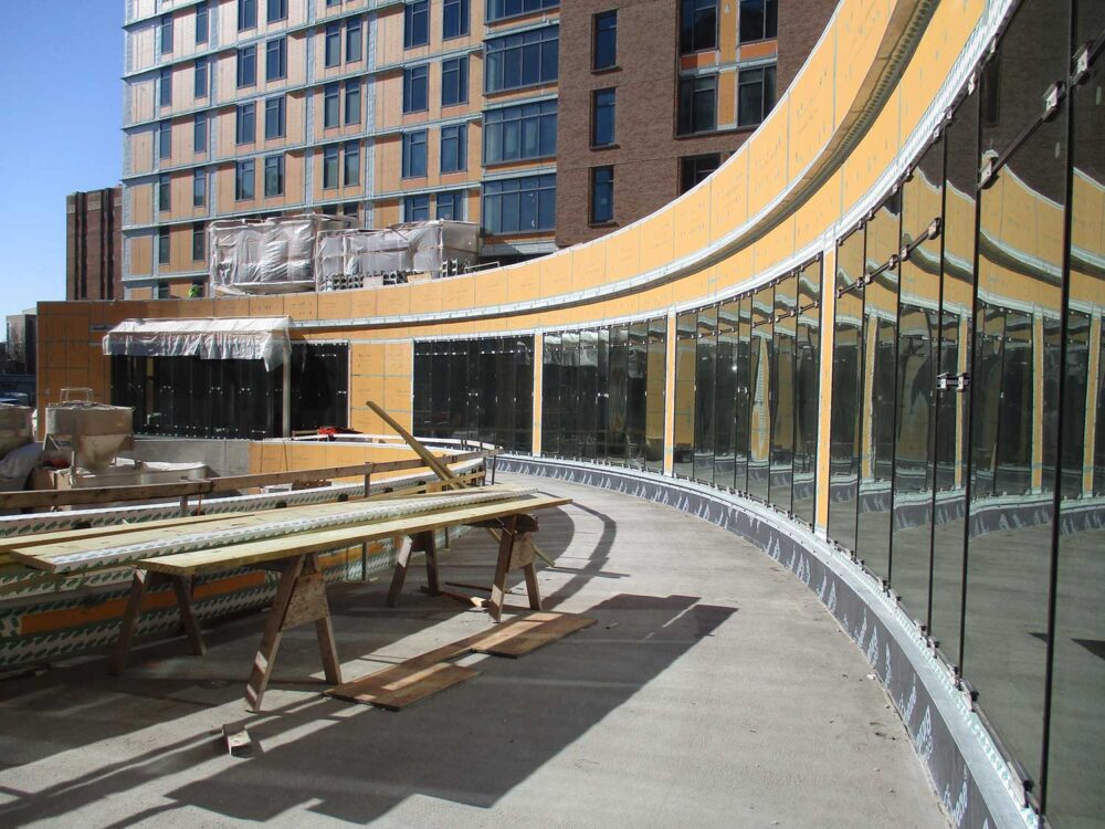 Curved glass wall under construction on a rooftop terrace, with yellow-sheathed walls, sawhorses and lumber in foreground.